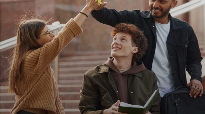 Drei junge Erwachsene sitzen auf einer Treppe im Freien, lachen und unterhalten sich. Eine Frau und ein Mann geben sich ein High-Five unter einem Baum, während der dritte im Sitzen ein Buch hält und lächelnd zu ihnen aufschaut. Die Szene wirkt freundlich und lebendig, typisch für einen Campus oder öffentlichen Platz.