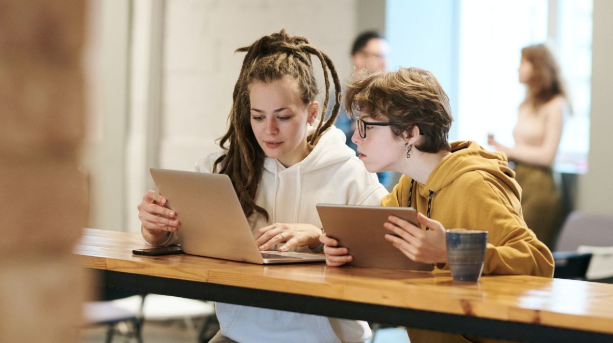 Zwei junge Personen arbeiten gemeinsam an einem langen Holztisch in einem modernen Arbeitsumfeld. Eine Person trägt einen weißen Hoodie und arbeitet an einem Laptop, die andere im gelben Hoodie hält ein Tablet. Beide sind konzentriert bei der Arbeit. Im Hintergrund unterhalten sich zwei weitere Personen. Die Szene wirkt kollaborativ und kreativ.