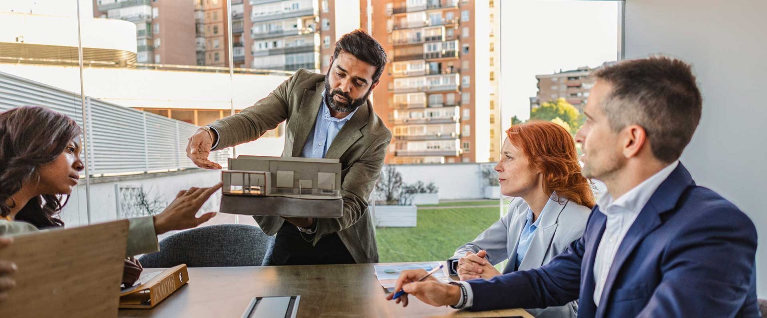 Personen im Besprechungsraum, mit Architekturmodell; im Hintergrund Fenster mit Stadtblick.