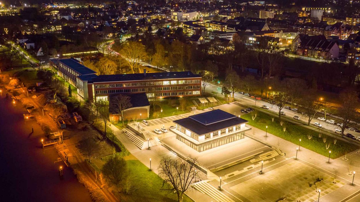 Beleuchteter Hochschulcampus bei Nacht mit moderner Architektur, zentralem quadratischen Gebäude und Blick auf Stadt und Fluss mit Schleusen.