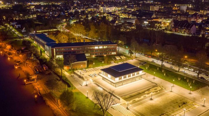 Beleuchteter Hochschulcampus bei Nacht mit moderner Architektur, zentralem quadratischen Gebäude und Blick auf Stadt und Fluss mit Schleusen.