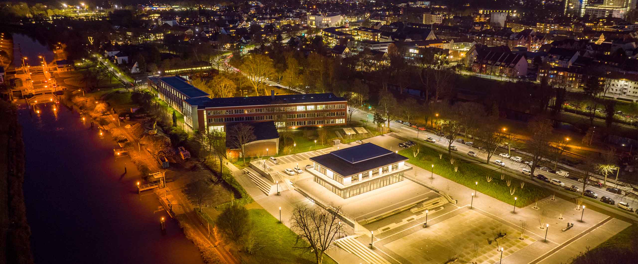 Beleuchteter Hochschulcampus bei Nacht mit moderner Architektur, zentralem quadratischen Gebäude und Blick auf Stadt und Fluss mit Schleusen.
