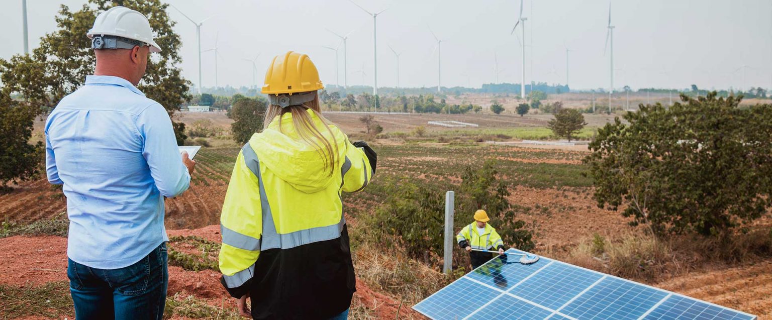 Drei Personen mit Warnwesten bei Solar- und Windanlagen; eine zeigt, eine notiert.