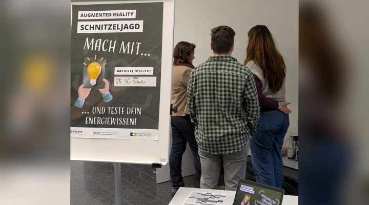 Ein Plakat auf einem Aufsteller wirbt für eine Augmented-Reality-Schnitzeljagd mit dem Text: ‚Mach mit … und teste dein Energiewissen!‘. Die aktuelle Bestzeit ist handschriftlich mit ‚05:40 Team Schollis‘ eingetragen. Im Hintergrund stehen drei Personen mit dem Rücken zur Kamera und schauen sich offenbar etwas an der Wand an. Auf einem Tisch im Vordergrund steht ein Tablet mit dem gleichen Plakatmotiv wie auf dem Aufsteller sowie ein ausgedruckter Plan oder Ablaufzettel.