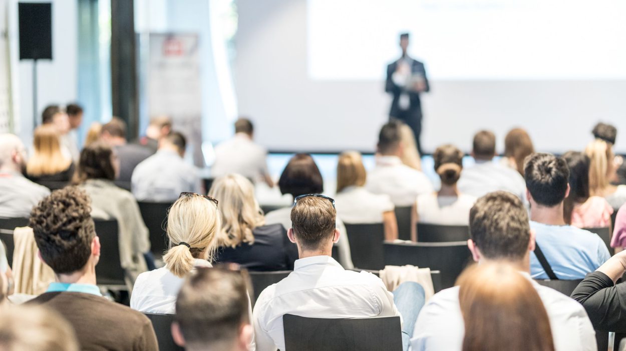 Ein Vortrag vor großem Publikum: Eine größere Gruppe von Menschen sitzt in Reihen in einem hellen, modernen Konferenzraum. Die Teilnehmenden blicken auf eine Bühne, auf der eine Person in Anzug und mit Mikrofon steht und präsentiert. Im Hintergrund ist eine große Projektionsfläche zu sehen, deren Inhalt unscharf ist. Die Atmosphäre wirkt professionell und konzentriert.