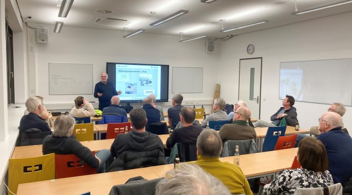 Ein Vortrag in einem Klassenzimmer: Eine Person steht vorne an der Tafel und präsentiert vor einer Gruppe von etwa 20 Zuhörenden, die an Tischen sitzen. Auf dem Bildschirm hinter dem Vortragenden ist eine Präsentationsfolie mit dem Titel ‚Wärmepumpencheck – Pilot‘ zu sehen. Die Teilnehmenden sind überwiegend älter, hören aufmerksam zu und einige trinken aus Glasflaschen. Der Raum ist hell beleuchtet und mit bunten Stühlen ausgestattet.