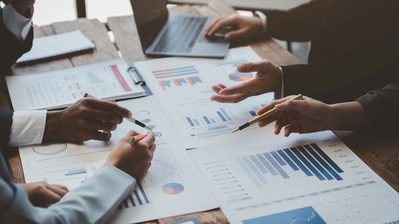 Gruppe analysiert Geschäftsdaten an Holztisch mit Diagrammen und Laptop.