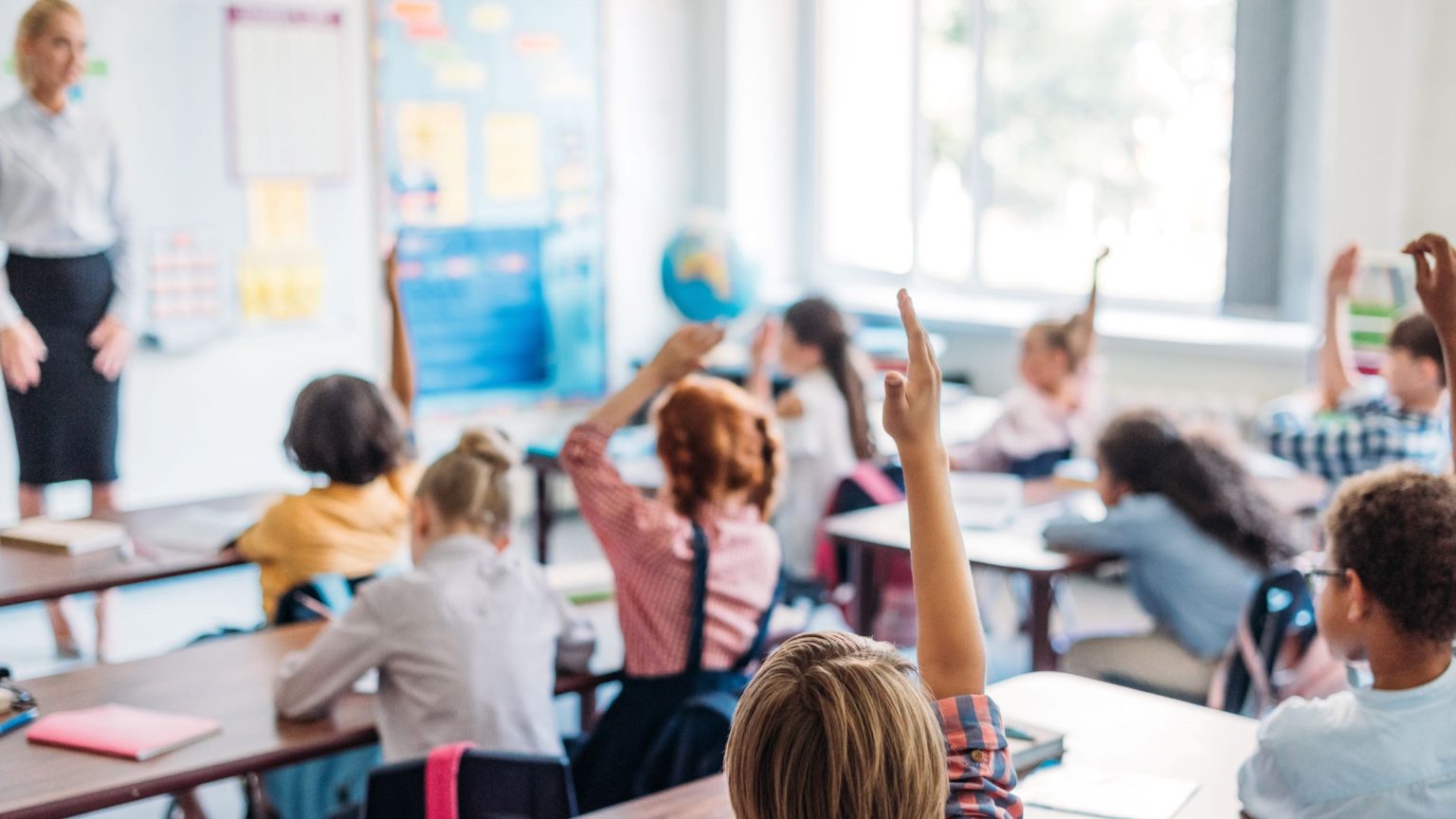 Grundschulkinder in hellem Klassenzimmer, mehrere melden sich; Lehrerin an der Tafel.