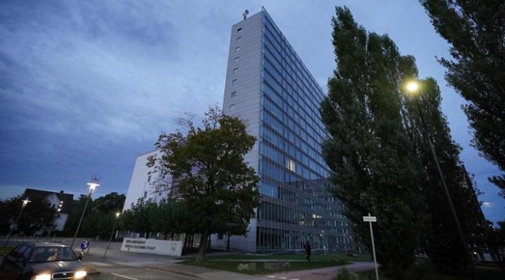 Ein modernes Hochhaus mit Glasfassade bei einsetzender Dämmerung. Das Gebäude steht an einer Straßenkreuzung, links fährt ein Auto vorbei. Im Vordergrund sind große Bäume und ein Schild mit der Aufschrift ‚Zentrum für Transformation und Nachhaltigkeit‘ zu sehen.