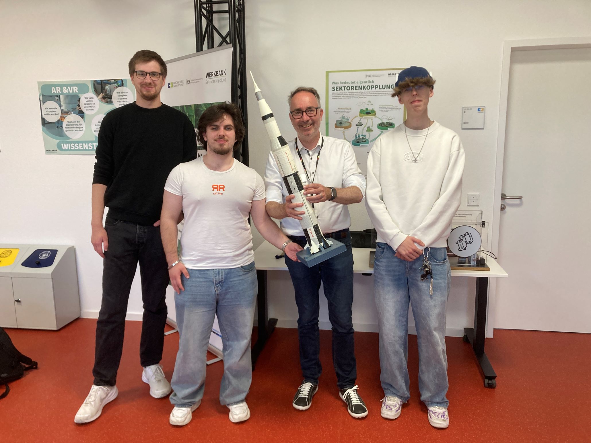Männer mit Saturn-V-Modell vor Postern zu Sektorenkopplung und AR/VR.